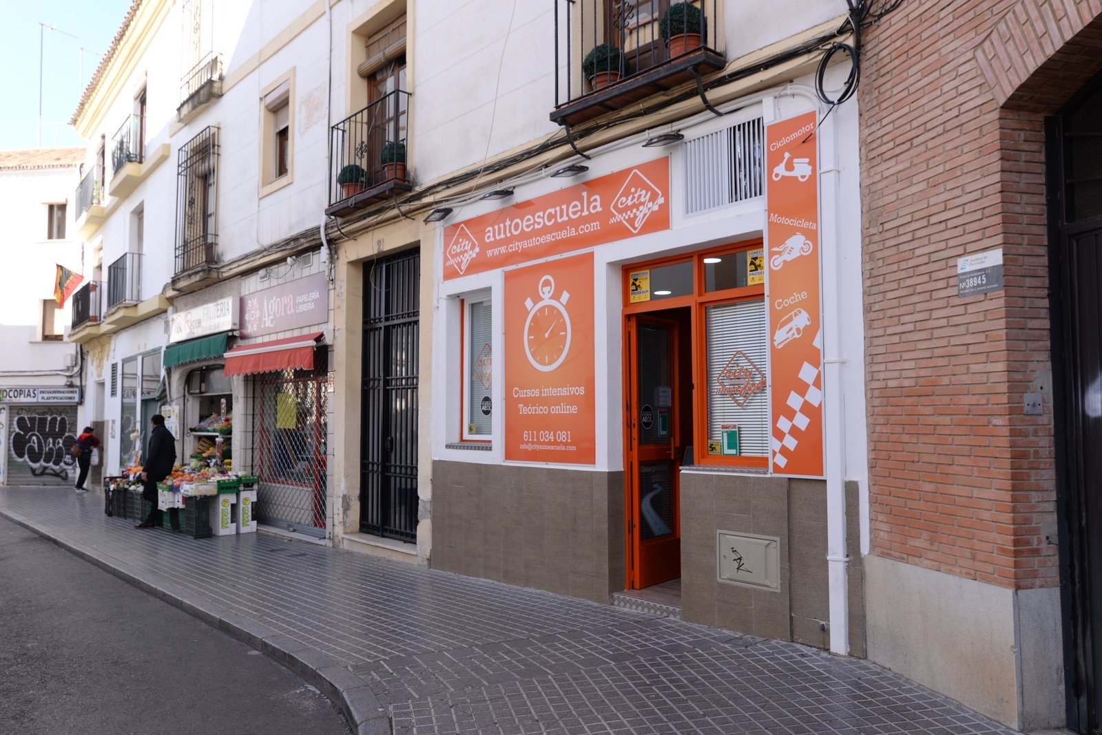 La fachada naranja de City Autoescuela vista desde la calle, junto a la frutería y la papelería del barrio