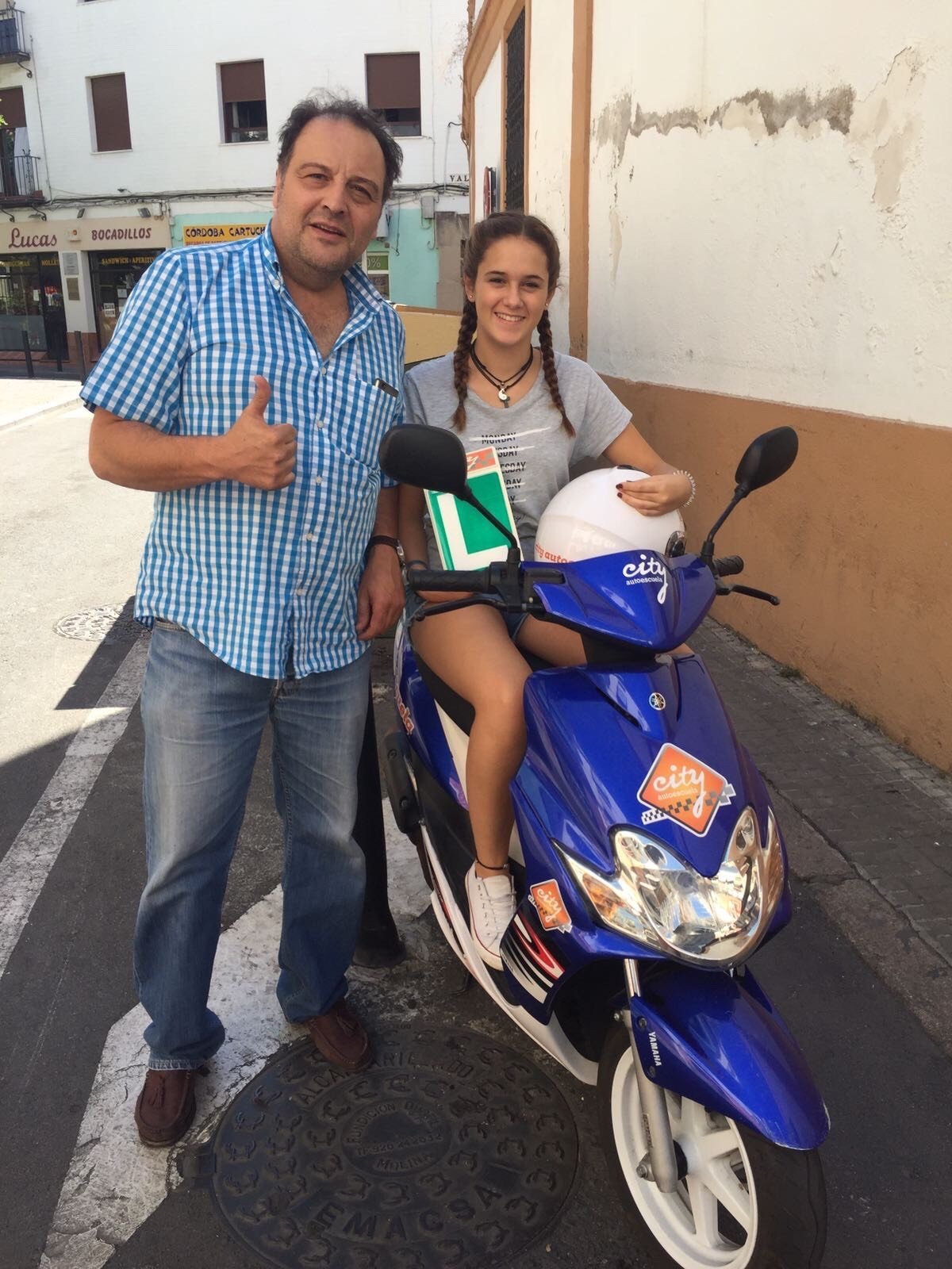 Profesor de City Autoescuela con el pulgar arriba junto a una alumna aprobada sobre el ciclomotor Yamaha Jog en la calle